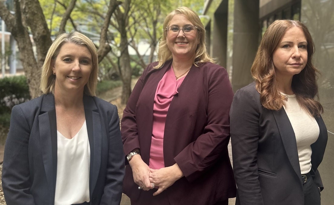 Three female solicitors standing outside a building, looking towards the camera, with green trees and shrubs in the background.