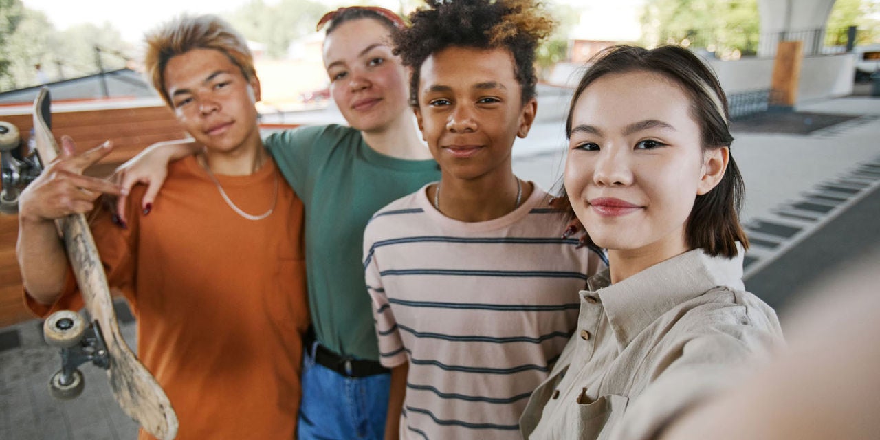 Four smiling young people taking a selfie together. The young person on the left holds a skateboard.