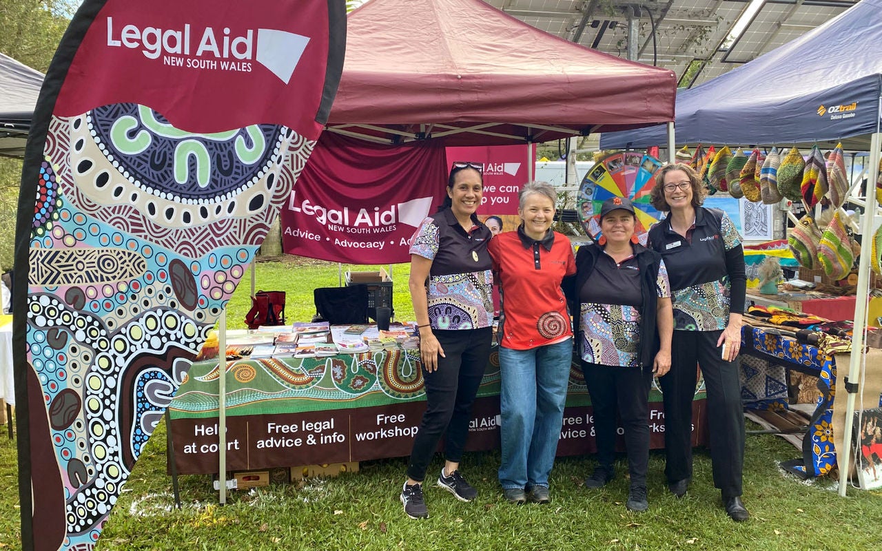 A photo of four people standing in front of a festival stall. In the foreground, there is a banner with the Legal Aid NSW logo and an artwork titled Lore and Law by Brungle artist Luke Penrith.