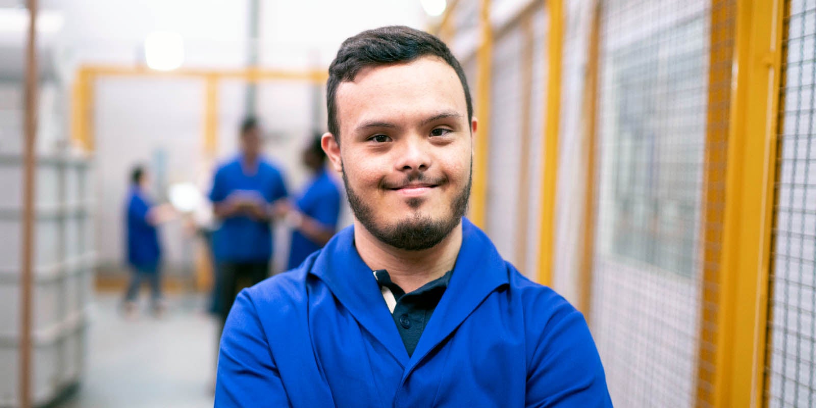 A worker in a blue uniform smiling at the camera. Other workers are in the background out of focus.