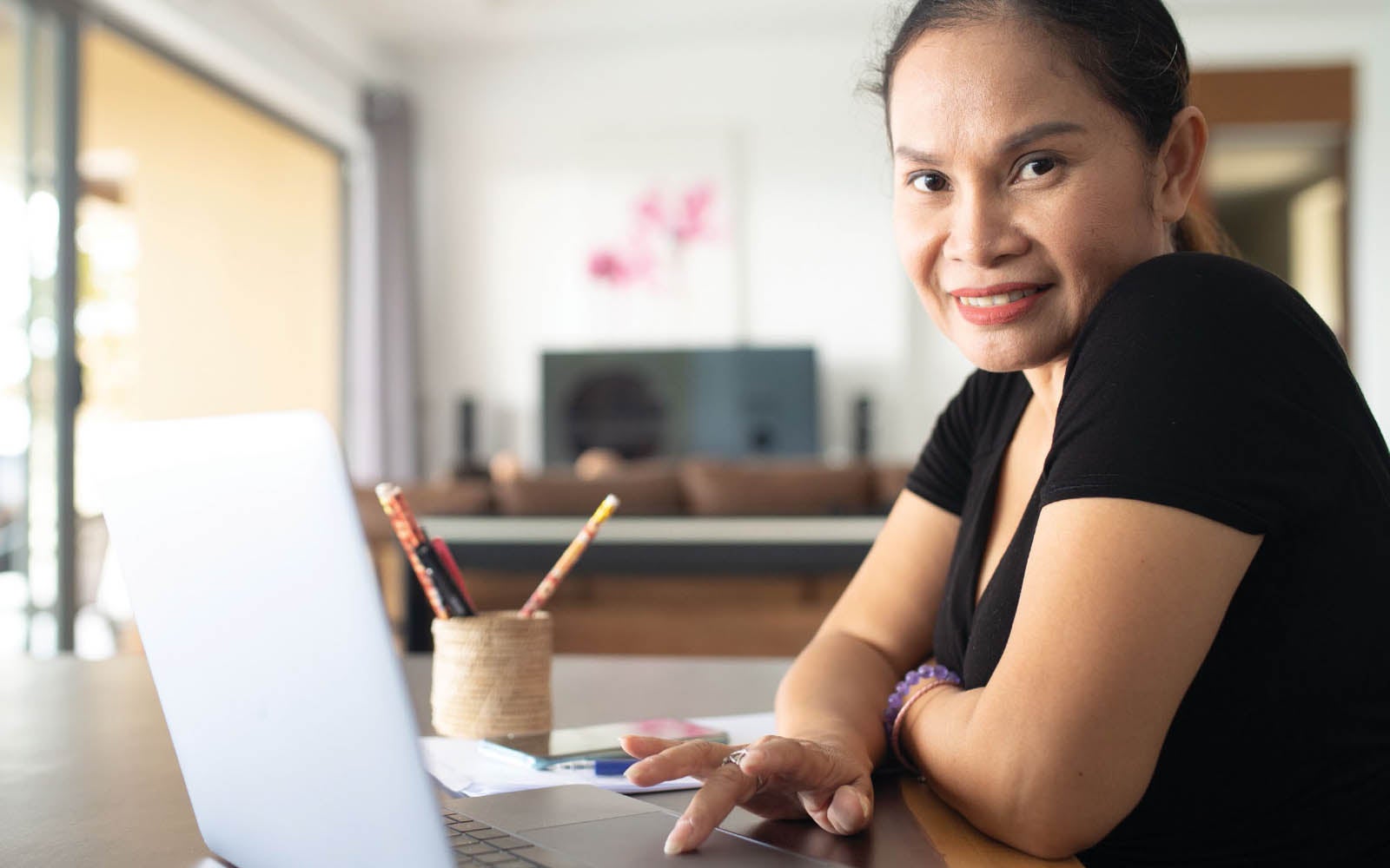 A smiling woman looking at the camera, sitting at a table and using the laptop in front of her.