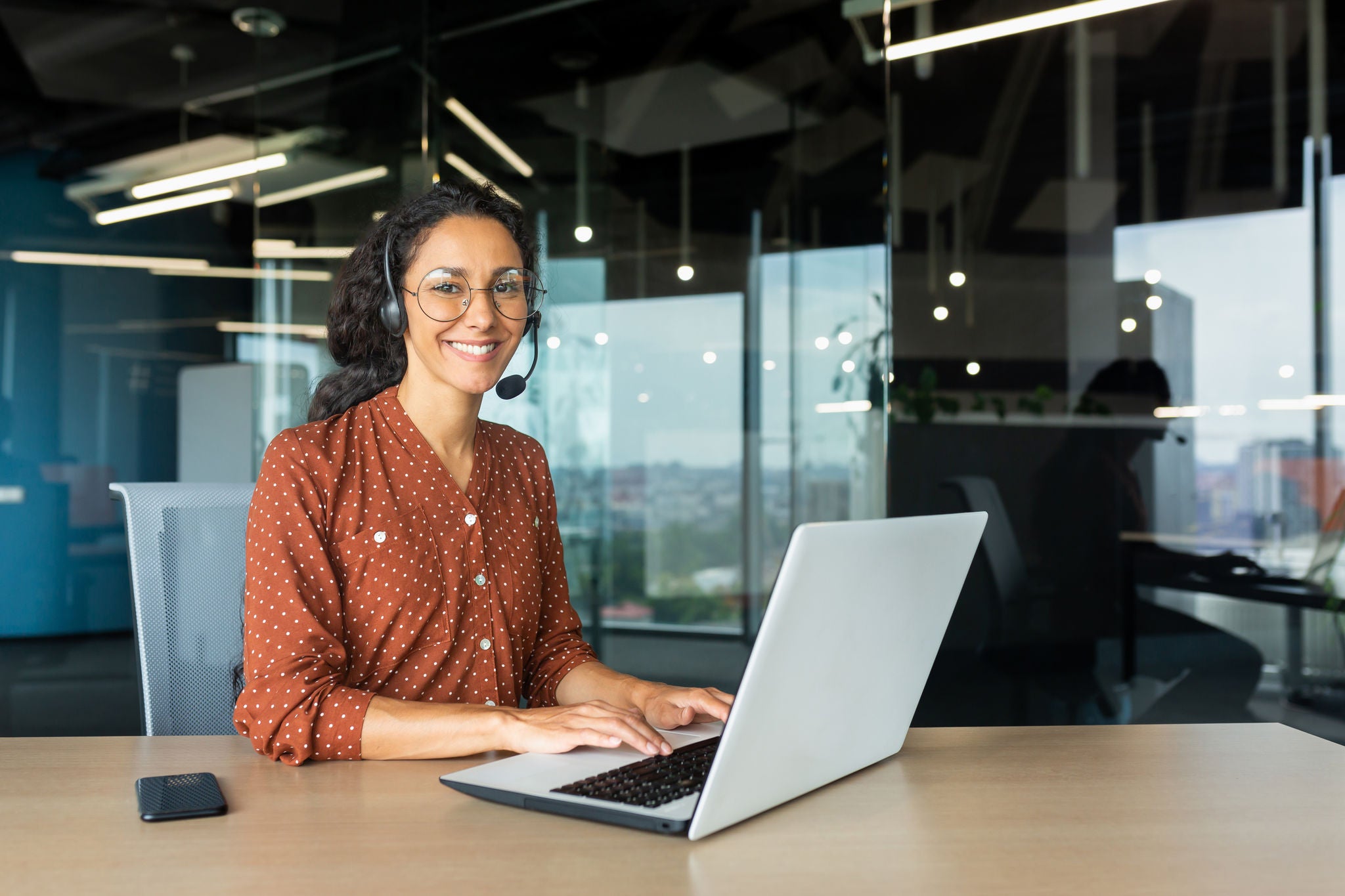 Portrait of business woman, office worker looking at camera and smiling, using headset and laptop for remote online communication, customer support tech call center worker.