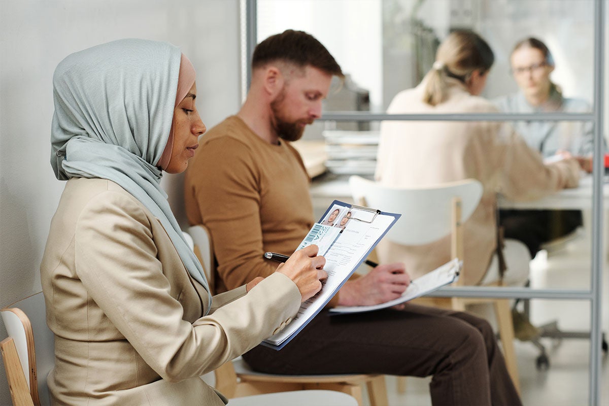 Man and woman sitting in room filling in form with two ladies on the background having a meeting