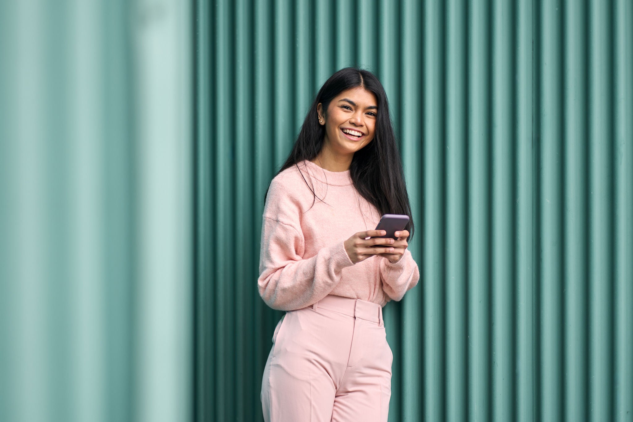 Happy Asian teen girl wearing pink outfit using mobile phone apps outdoors looking at camera. Positive smiling pretty female model holding smartphone laughing standing at blue wall background.