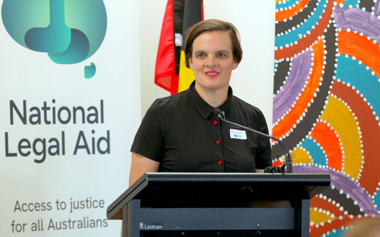 Katherine stands at a lectern smiling. Behind her is a banner for National Legal Aid and a bright and colourful painting.