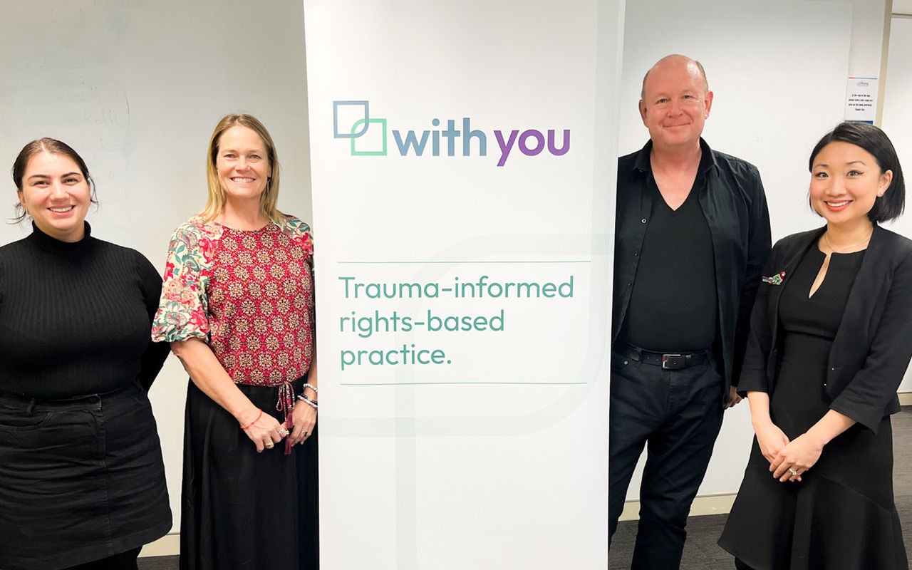 A photo of four people in an office standing next to a banner. On the left is Kate Adnams and Arna Rathgen. In the centre the banner reads ‘with you – trauma-informed rights-based practice.’ On the right is Darren Wagner and Jennifer Chen.