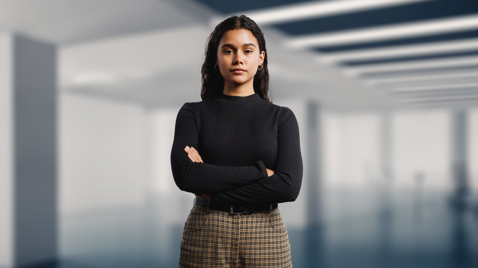 A young woman stands in the foreground of an office building. She has her arms folded.