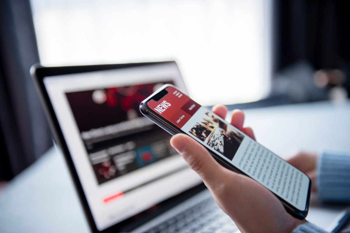 A hand holding a mobile phone that displays a news article is visible in the foreground. Behind the mobile phone a laptop is open but the screen is out of focus.