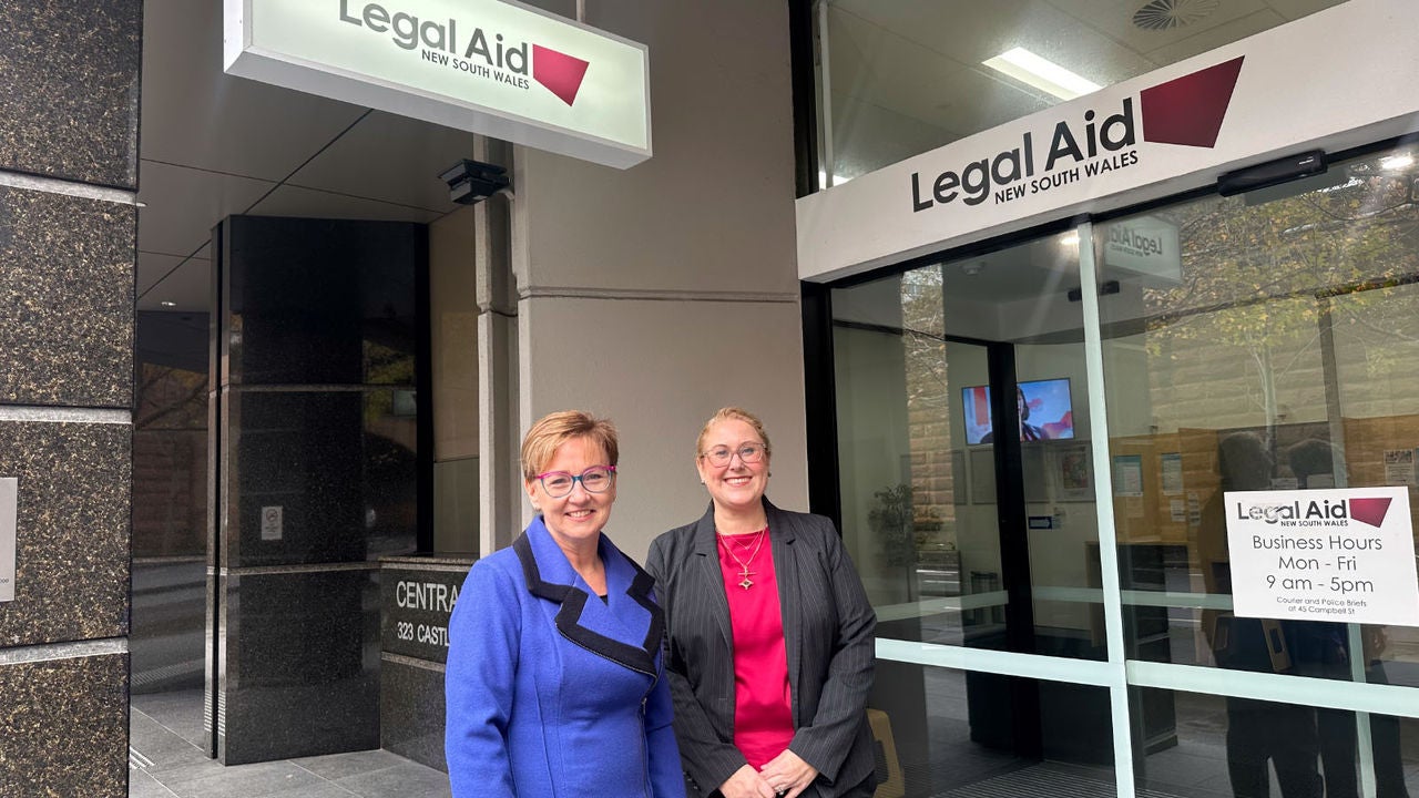 Jodie and Anna stand in front of a Legal Aid NSW office. Legal Aid NSW signs are visible behind them. Jodie is wearing a bright blue coat and Anna has a bright red top underneath a striped jacket. Both women are smiling.